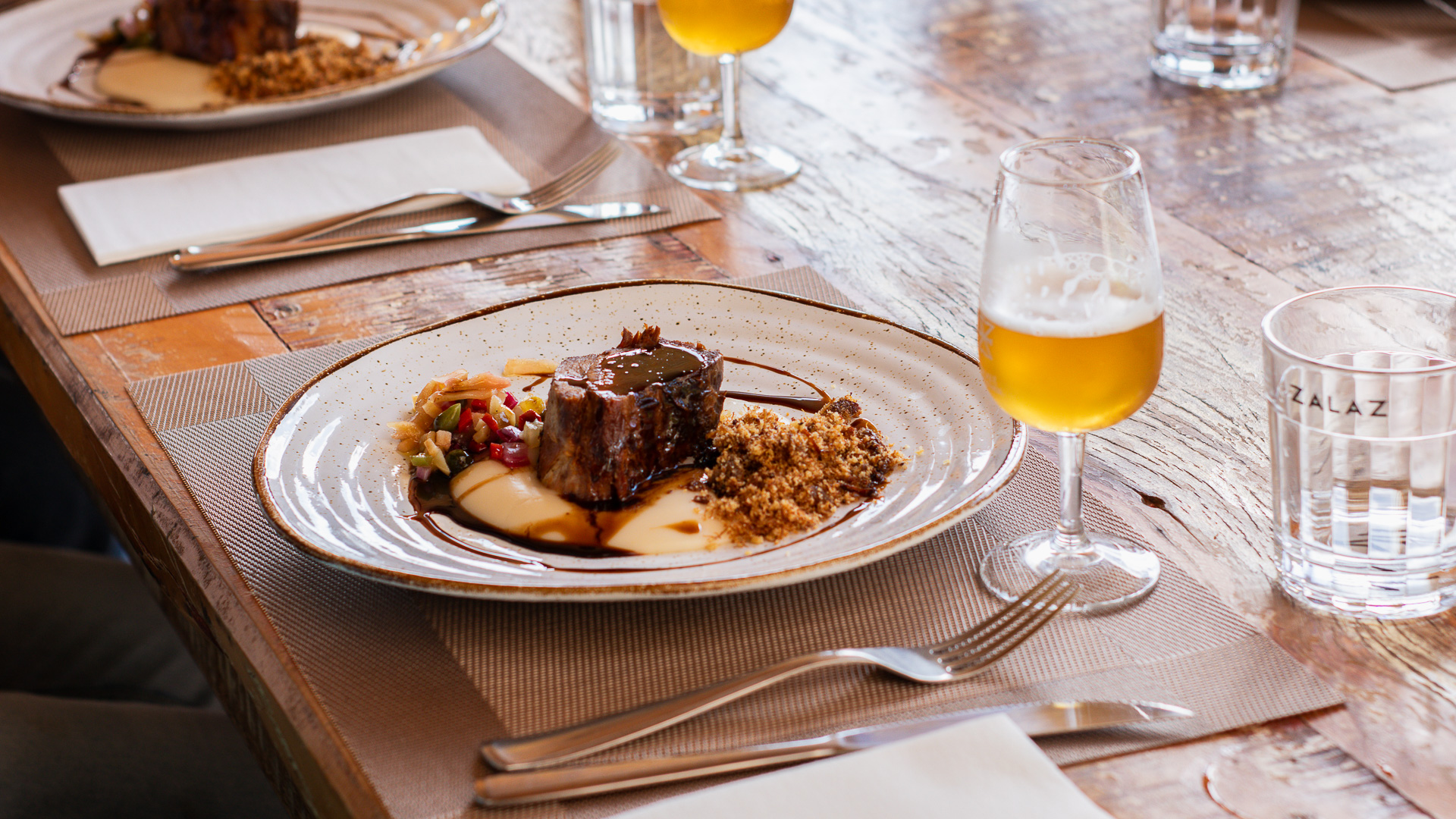 Prato gourmet da ZalaZ: carne bovina braseada com purê cremoso e farofa, acompanhada de cerveja artesanal dourada e copo com a logo ZALAZ, sobre mesa de madeira rústica